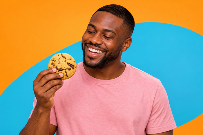 Lachender Mann mit Chocolate Chip Cookie vor orangefarbenem und türkisfarbenem Hintergrund